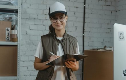 pexels-photo-6170101-6170101 Smiling woman holding clipboard in office, managing logistics with eco-friendly focus.
