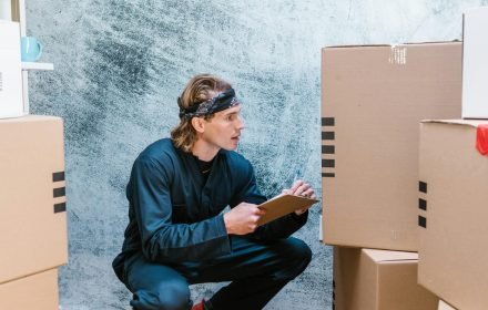 pexels-photo-7464380-7464380 A man crouches to check shipping boxes, clipboard in hand, indoors.